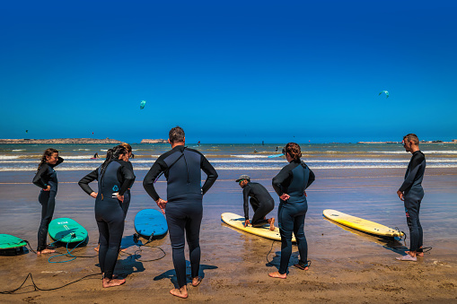 Essaouira, Morocco - August 3, 2023: A group of young people in wetsuits on the beach listen to lesson from an expert who teaches them the sport of kitesurfing.