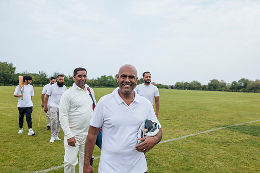 Male friends and family members heading out into a field to play cricket together. They are wearing white, carrying their equipment on a sunny day in Northumberland. Some are smiling looking at the camera.