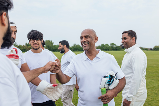 Male friends and family members heading out into a field to play cricket together. They are wearing white, fist bumping/greeting each other.