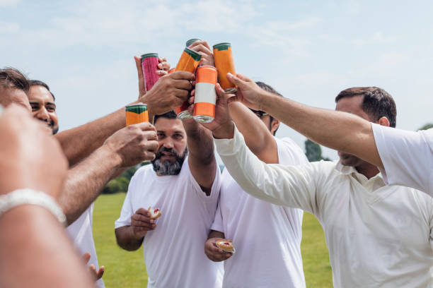 Male friends and family members celebrating together after playing cricket together. They are wearing white standing with their hands together, toasting with their soft drinks in a huddle on a sunny day in Northumberland.