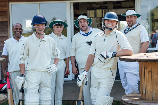 Male cricket team ready to play a game together on a sunny day in Northumberland. They are standing outside of their clubhouse wearing sports helmets, laughing together.
