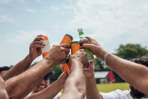 Male friends and family members celebrating together after playing cricket together. They are wearing white standing with their hands together, toasting with their soft drinks in a huddle on a sunny day in Northumberland. Focus on the drinks.