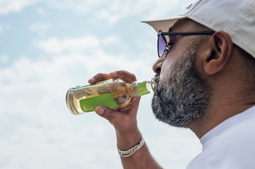 Close up of a man drinking from a bottle outdoors in  the sunshine in Northumberland. He is wearing a cap and sunglasses.