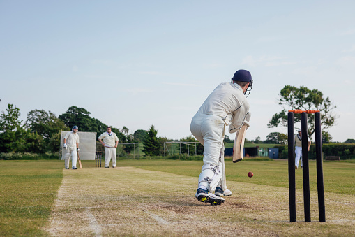 Male cricket team members playing a game together. They are wearing white enjoying the game on a sunny day in Northumberland. The men are all at their posts, batting, keeping and catching. Focus on the man batting.
