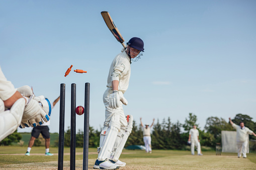 Male cricket team members playing a game together. They are wearing white enjoying the game on a sunny day in Northumberland. The men are all at their posts, batting, keeping and catching. Focus is on the cricket pegs are in mid air.