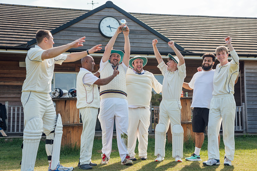 Male cricket team celebrating winning a game together on a sunny day in Northumberland. They are standing outside of their clubhouse laughing together, cheering holding a trophy.