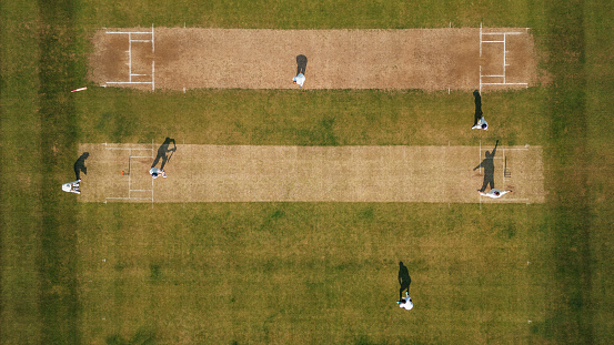 Aerial view of a male cricket team playing a game together. They are wearing white enjoying the game on a sunny day in Northumberland. The men are all at their posts, batting, keeping and catching.