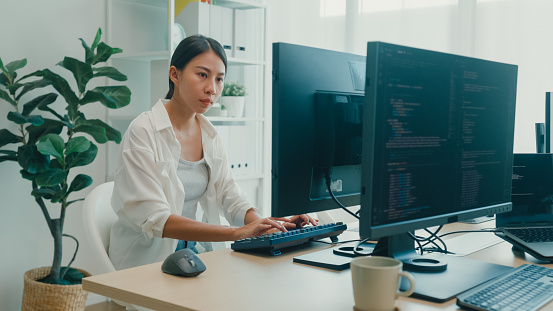 Young Asian woman software developers using computer to write code sitting at desk with multiple screens work at office. Programmer development concept.