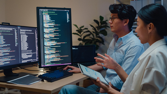Group of Asian people software developers using computer to write code sitting at desk with multiple screens in office at night. Programmer development concept.