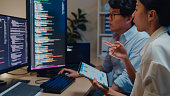 Group of Asian people software developers using computer to write code sitting at desk with multiple screens in office at night. Programmer development.