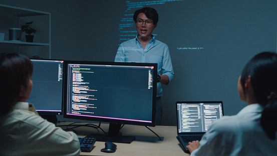 Young Asian man software developers mentor leader manager talking to executive team analyzing source code in office at night. Programmer development concept.