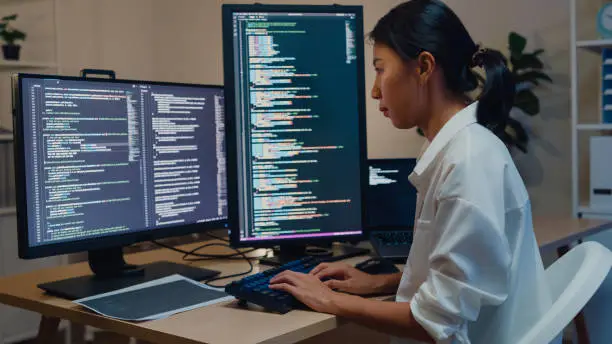 Photo of Young Asian woman software developers using computer to write code sitting at desk with multiple screens work in office at night. Programmer development.