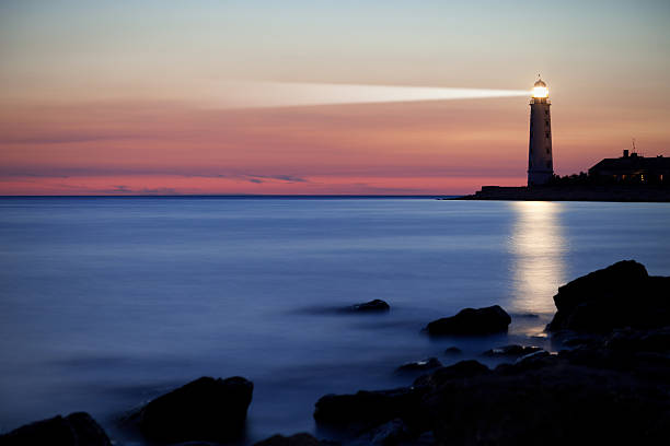 A lighthouse on the coast at sunset A seascape is pictured at the end of sunset. There is a lighthouse casting light into the background and reflecting onto the waters in front of it. In the foreground there are silhouetted rocks, in the middle ground there is soft blue-white water and in the background is the lighthouse and colorful sky filled with pink, orange and green. Beacon stock pictures, royalty-free photos & images