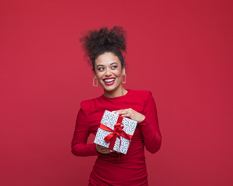 Young woman wearing red elegant dress, holding Christmas present, looking away and laughing. Studio shot, red background.