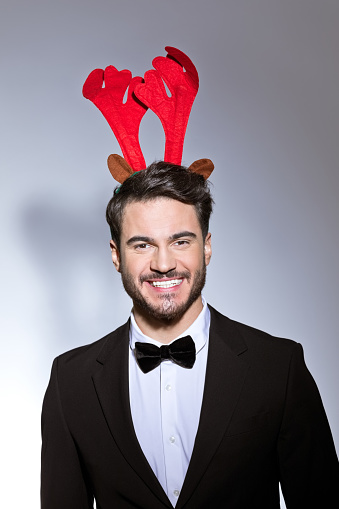 Excited young man wearing black elegant suit and red reindeer horns headband laughing at camera. Studio shot, white background.