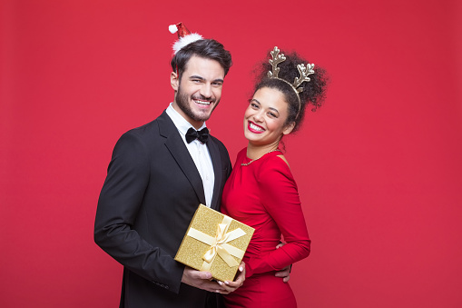 Excited young woman and man wearing elegant clothes and Christmas headbands embracing and smiling at camera. Man holding Christmas present. Studio shot, red background.