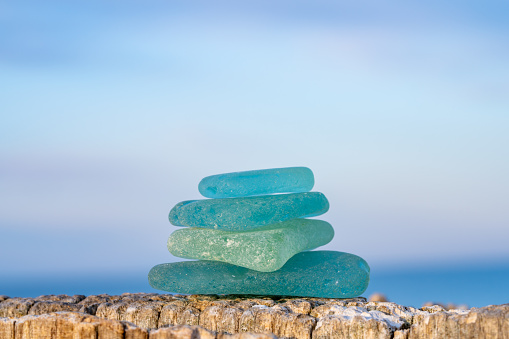 A stack of sea glass in blues and greens sits atop a piece of driftwood at the beach.