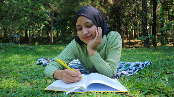 Relaxed muslim woman enjoying weekend at park, lying down on grass and writing book, empty space