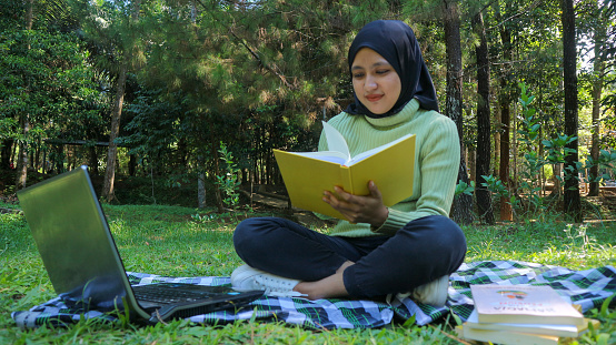 Relaxed muslim woman enjoying weekend at park, sitting on grass and reading book, empty space