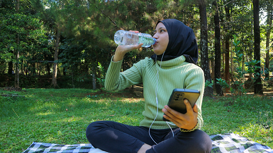 Happy muslim woman listen music on mobile phone at park while drinking mineral water, free space