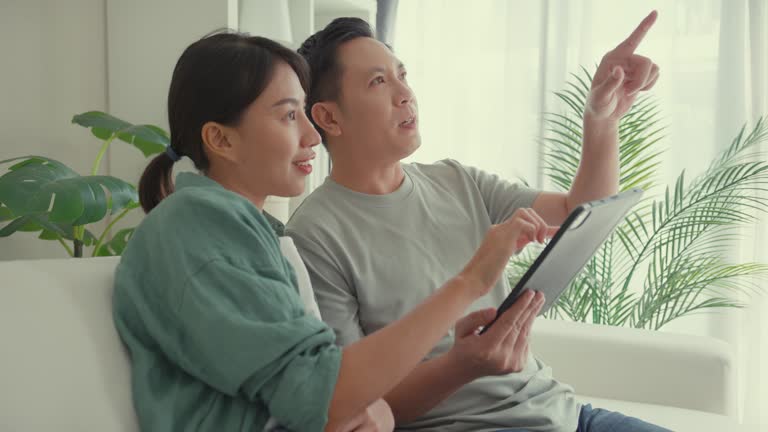 Closeup of happy Asian couple having fun using digital tablet devices online shopping together sit on sofa in living room at home. Lifestyle spend time in house.