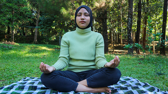 Portrait of peaceful muslim woman training and meditation with closed eyes at park. Harmony with nature and relaxation. Practicing of yoga.