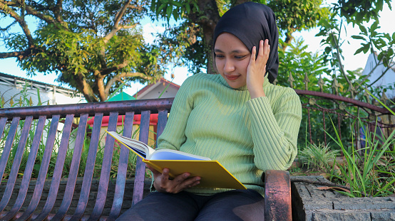 Relaxed muslim woman enjoying weekend at park, sitting on bench and reading book, empty space