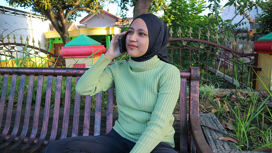 Happy asian muslim woman talking on mobile phone in park, free space