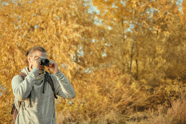 istock Man with a backpack looks into the camera viewfinder. Autumn time, trees with yellowed foliage. Copy space. 1646658086