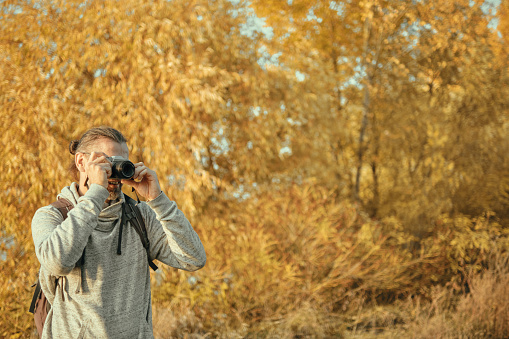 Man with a backpack looks into the camera viewfinder. Autumn time, trees with yellowed foliage. Copy space.