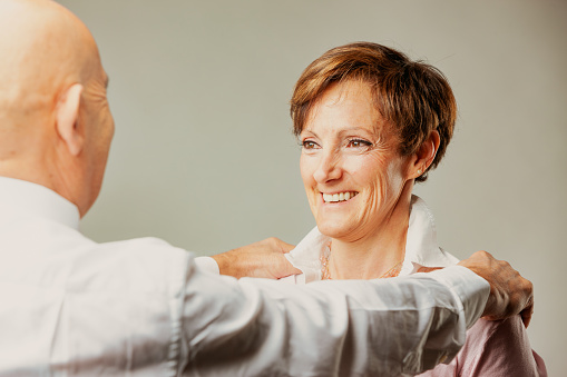 A bald man rests his hands on the shoulders of an elderly woman who laughs happily. He has a white shirt and she has a white blouse and pink sweater and perfect teeth.