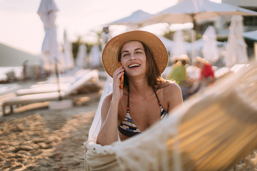 Beautiful young woman wearing a bikini using a smartphone and relaxing in a hammock chair at the beach and enjoying her summer vacation at the seaside