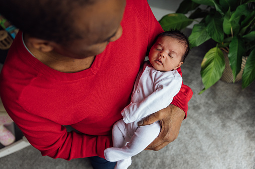 A senior man holding his granddaughter in his arms while standing in the living room in his house in Gateshead, North East England. He is looking and admiring the baby while she sleeps.