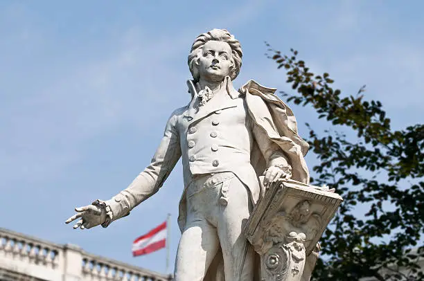 Statue of Wolfgang Amadeus Mozart, located in the Burggarten in Vienna. In the background the Austrian flag on the Austrian National Library. Selective Focus