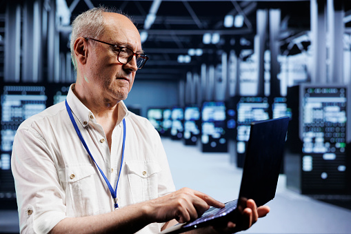 Senior IT developer inspects supercomputers in data center, ensuring smooth performance. Capable programmer in server farm monitoring energy consumption across units components using laptop