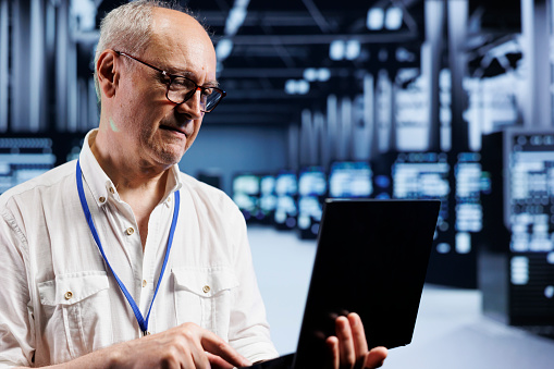 Senescent system administrator inspects server clusters in data center, ensuring smooth performance. Precise professional monitoring energy consumption across units components using laptop