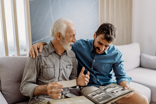 Close up portrait of grey haired he his him grandpa with daily newspaper widely toothy smiling laugh about political news wearing casual checkered shirt jeans outfit sitting comfy on divan
