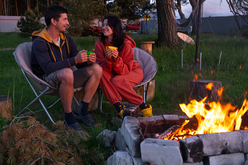 Couple in love is sitting by the outdoor campfire in the courtyard of the house on camping chairs, a romantic evening. a man and a woman warm their hands by the fire, drink tea