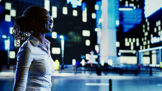 Smiling african american woman wandering around city boulevards during nighttime, pointing at interesting OOH advertisements on buildings. Businesswoman strolling around empty streets at night
