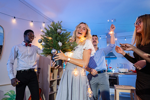 Young woman holding Christmas tree, while having fun during Christmas house party with her multiracial group of friends