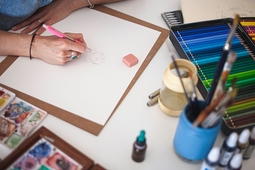 Table where illustrator works full of material and her hands working. You can see brushes, watercolors, colored pencils,...