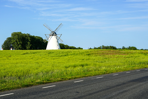 Historical white windmill in a green meadow on the side of the black asphalt road, blue sky background