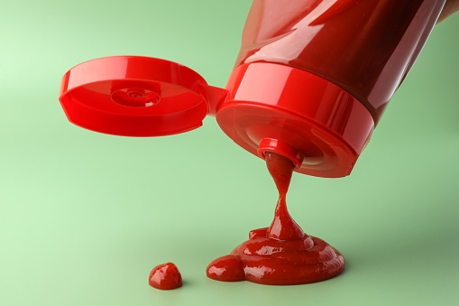 Pouring tasty red ketchup from bottle on green background, closeup