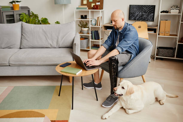 istock Man with prosthetic leg using laptop at home and petting labrador dog 1646903495