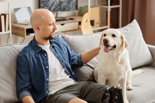 Portrait of smiling adult man with prosthetic leg relaxing on couch with happy white dog