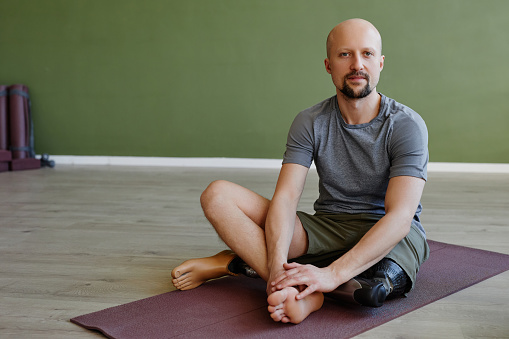 Full length portrait of adult man with prosthetic leg sitting on yoga mat indoors and looking at camera