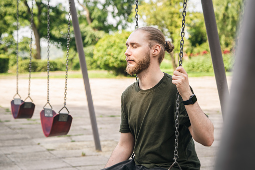 A young man with a beard is resting in the park, swinging on a swing, a man on the playground.