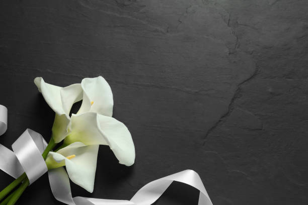 istock Beautiful calla lily flowers and white ribbon on black table, flat lay with space for text. Funeral symbols 1646908959