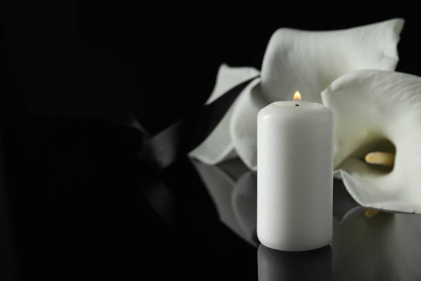 Burning candle and white calla lily flowers on black mirror surface in darkness, closeup with space for text. Funeral symbol