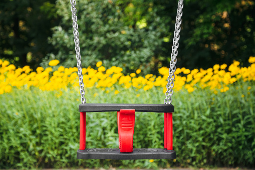 Red plastic swing hanging from chains in a children's park on a green blurred background.